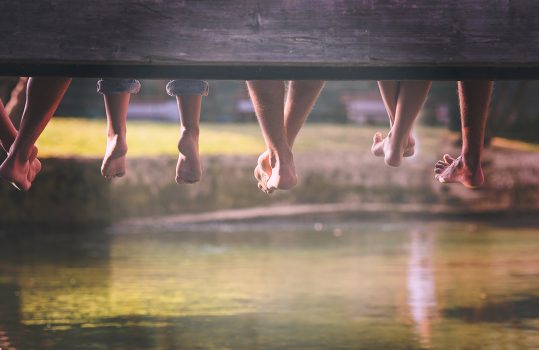 group of  people sitting at wooden bridge over the river with a focus on hanging legs